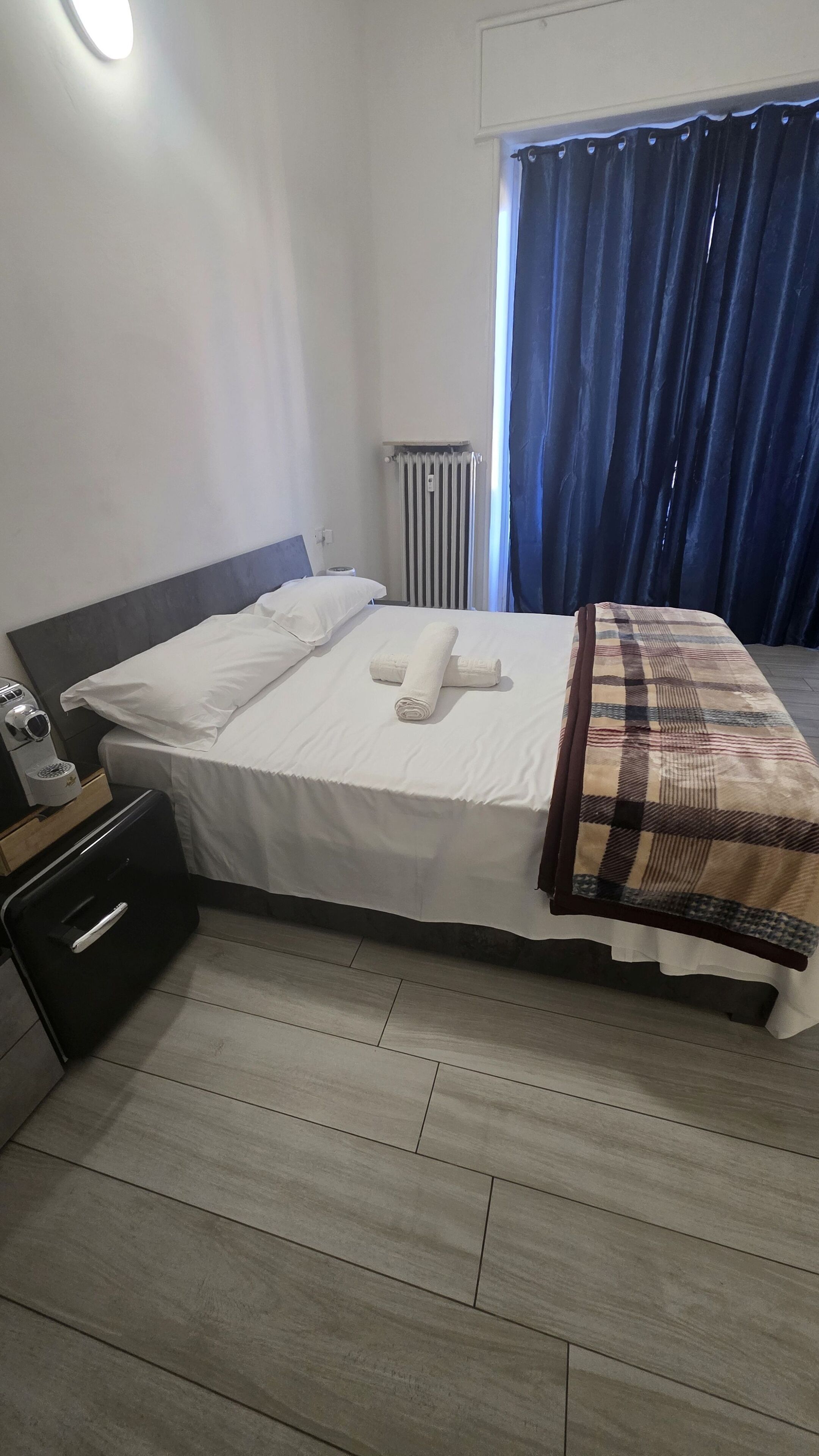 Une chambre d’hôtel avec un lit, une table de chevet avec une machine à café, un radiateur et une fenêtre avec des rideaux.