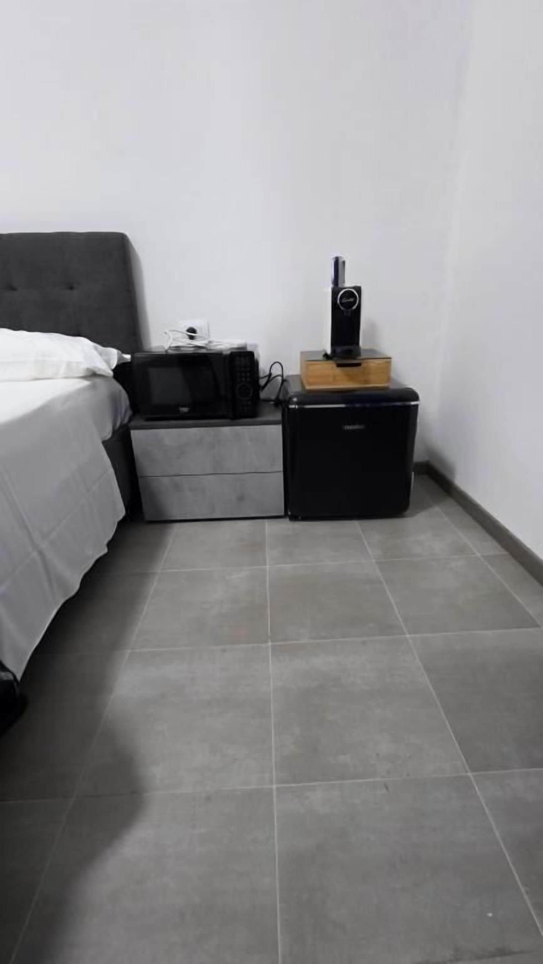 Un dormitorio con una cama, una mesita de noche, un pequeño electrodoméstico negro y un altavoz sobre un caja de madera.