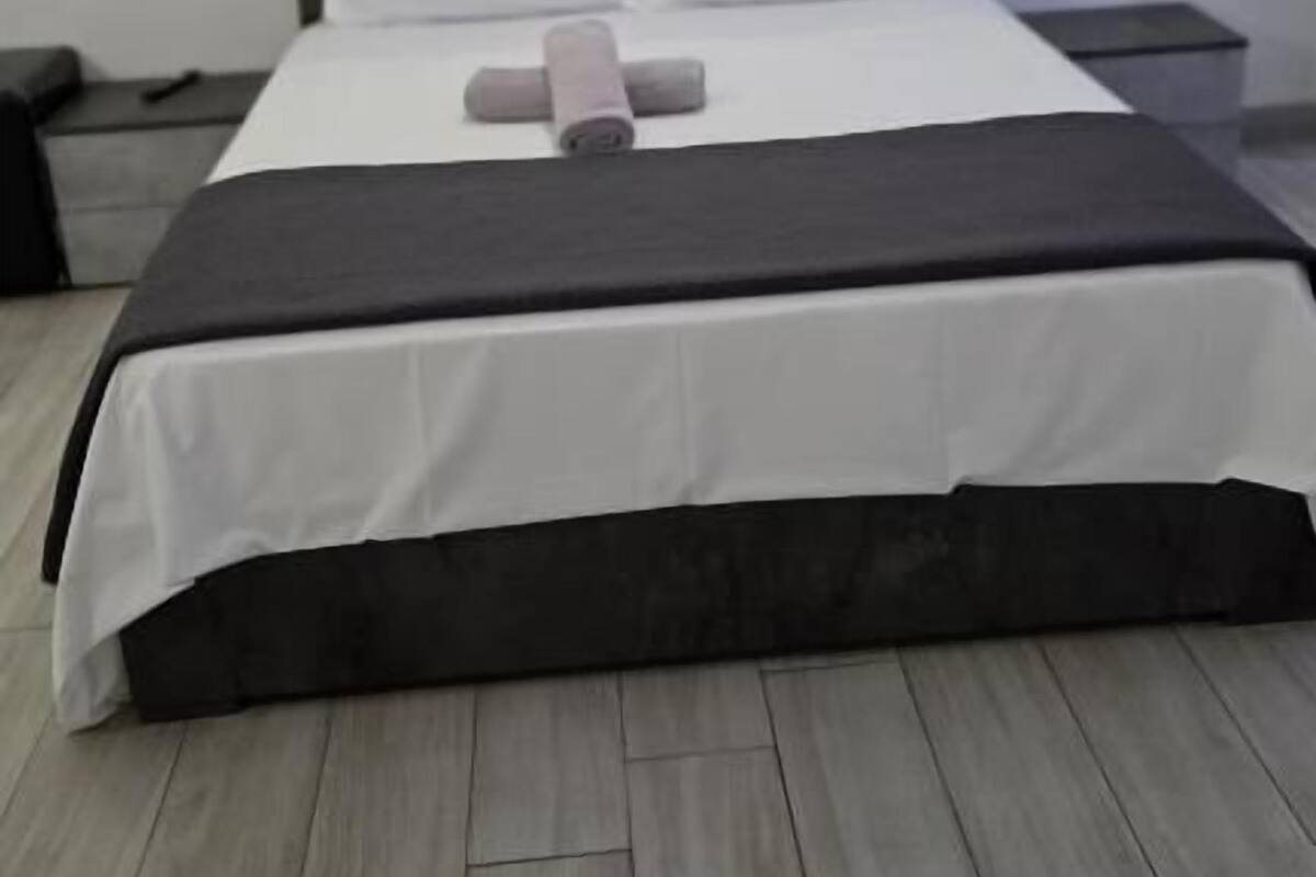 A neatly made bed with a white and black bedspread, a pillow, and a rolled-up towel. There is a nightstand with a lamp on it, and the room has a wooden floor.