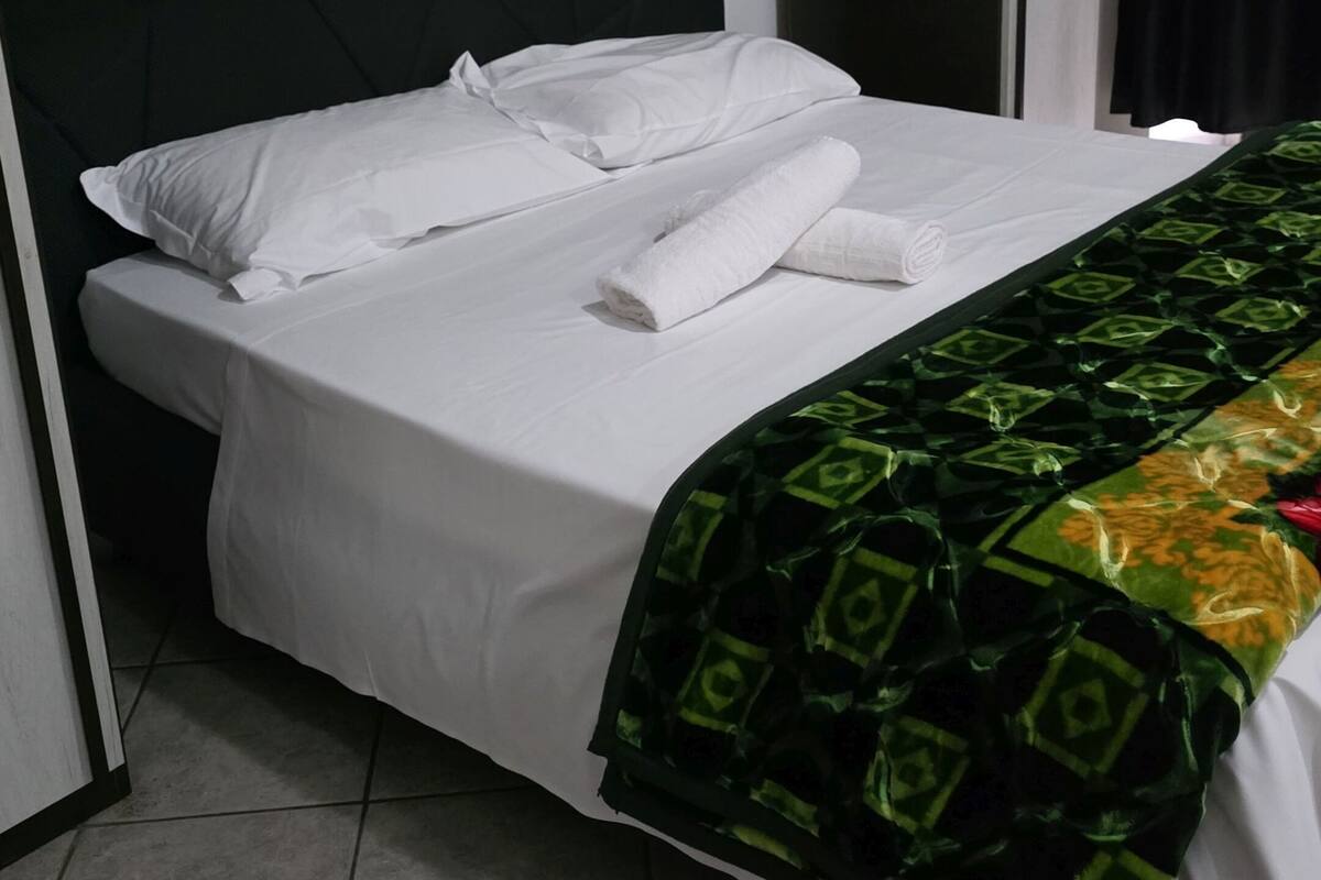 A neatly made bed with white linens and a green patterned blanket, a dark headboard, and light-colored wooden cabinets above.