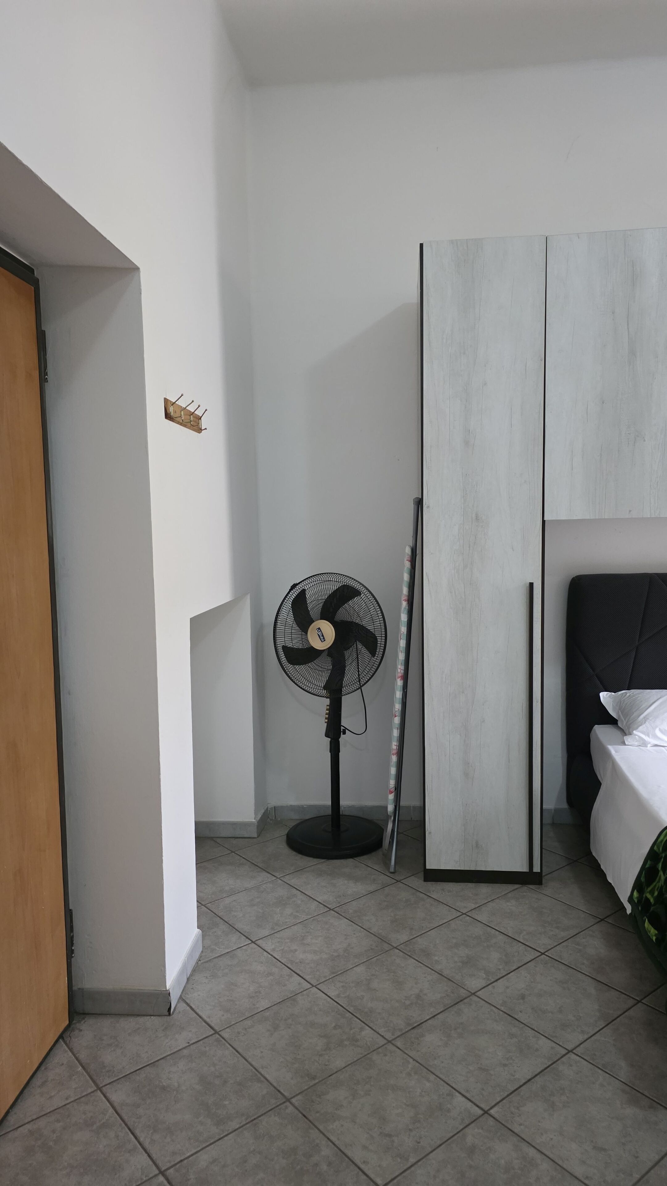 Ein Zimmer mit einem Bett, einem Standventilator, einer Holztür und einer weißen Wand mit einer kleinen Ablagefläche.