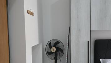 Ein Zimmer mit einem Bett, einem Standventilator, einer Holztür und einer weißen Wand mit einer kleinen Ablagefläche.