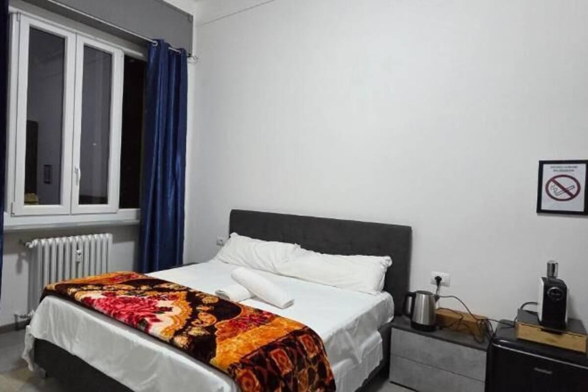 A bedroom with a bed, a window with curtains, a radiator, a nightstand with a coffee maker, and a trash bin.