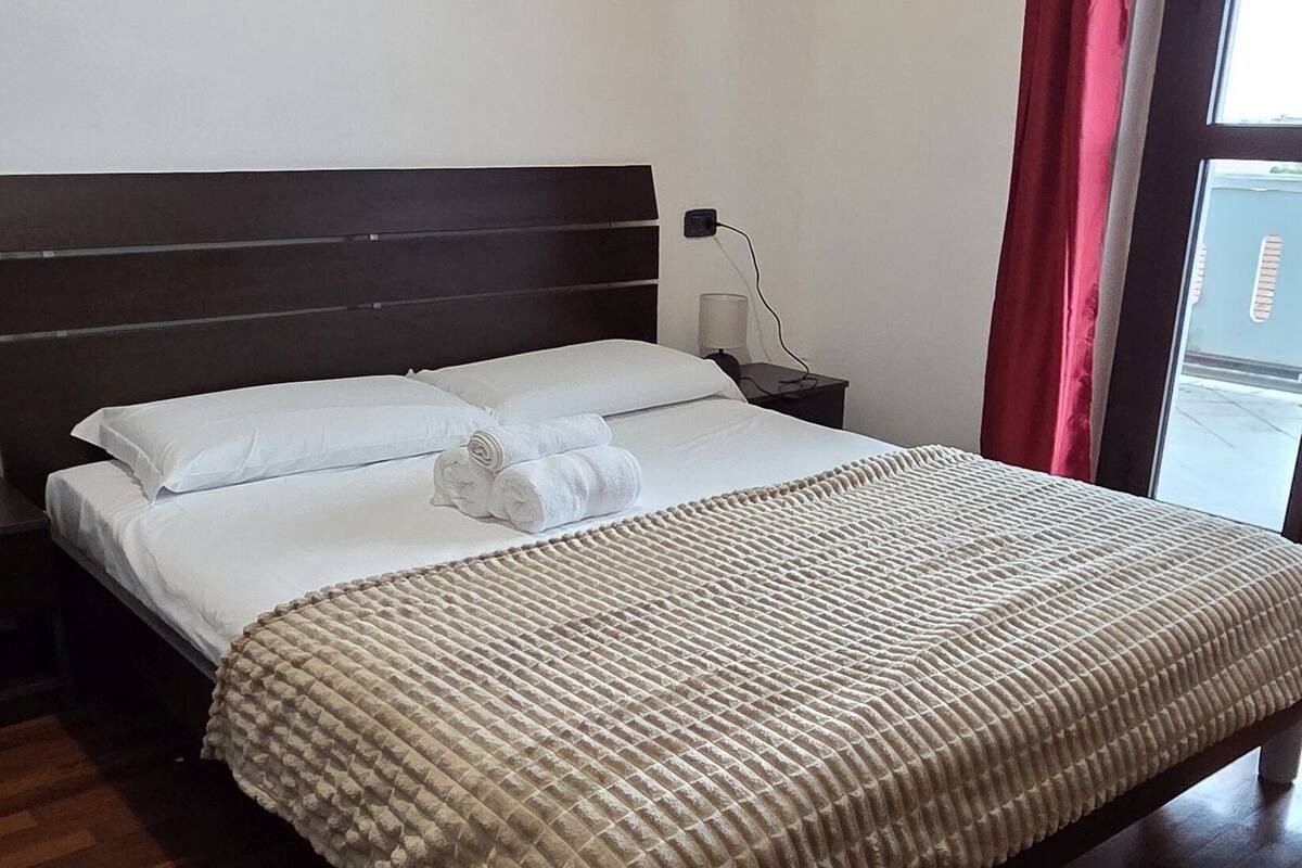 A single bed with a headboard, a bedside table, and a window with red curtains.