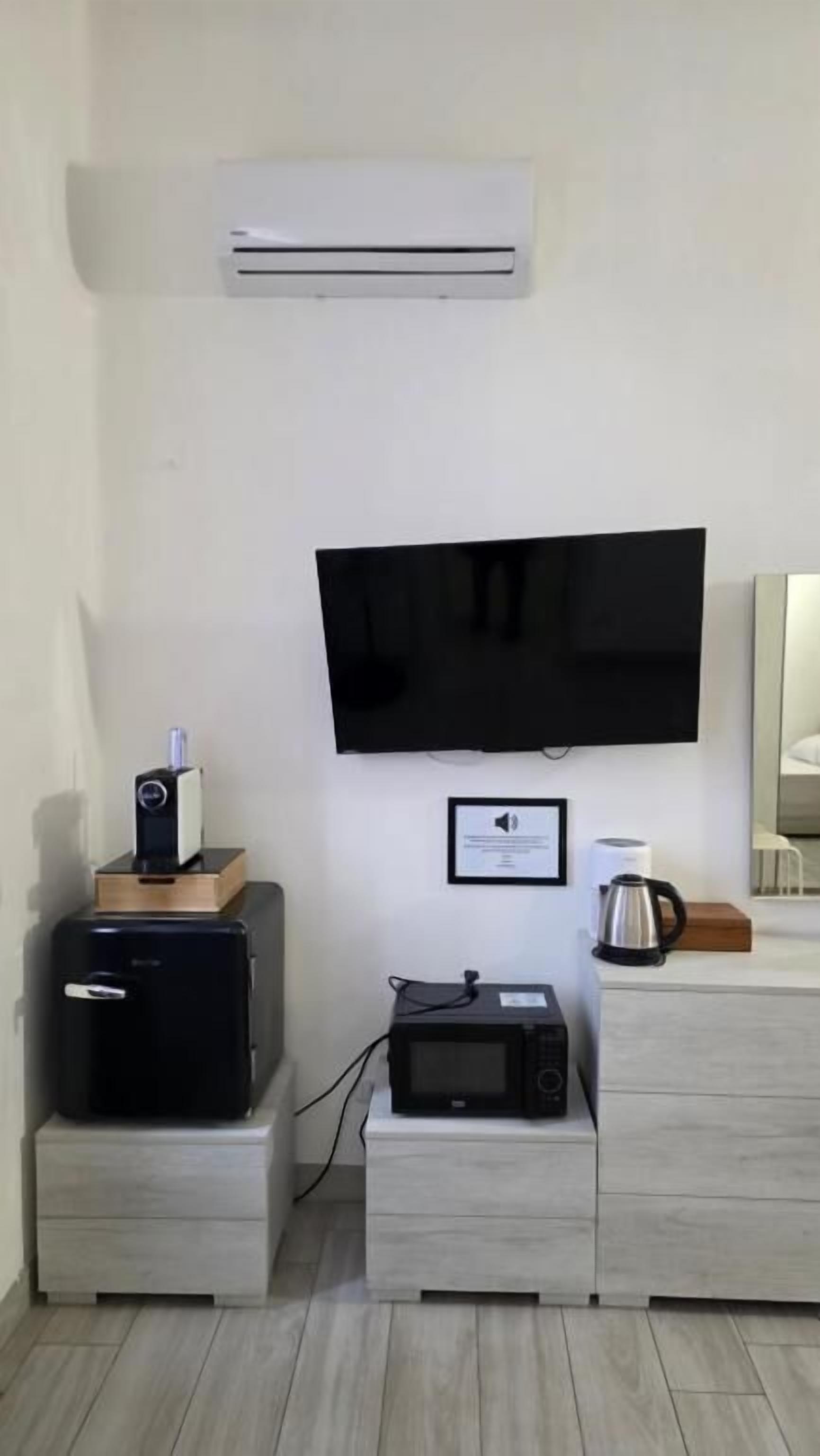 A room with a flat-screen TV mounted on the wall, a small refrigerator, a microwave, and a coffee maker on a wooden cabinet.