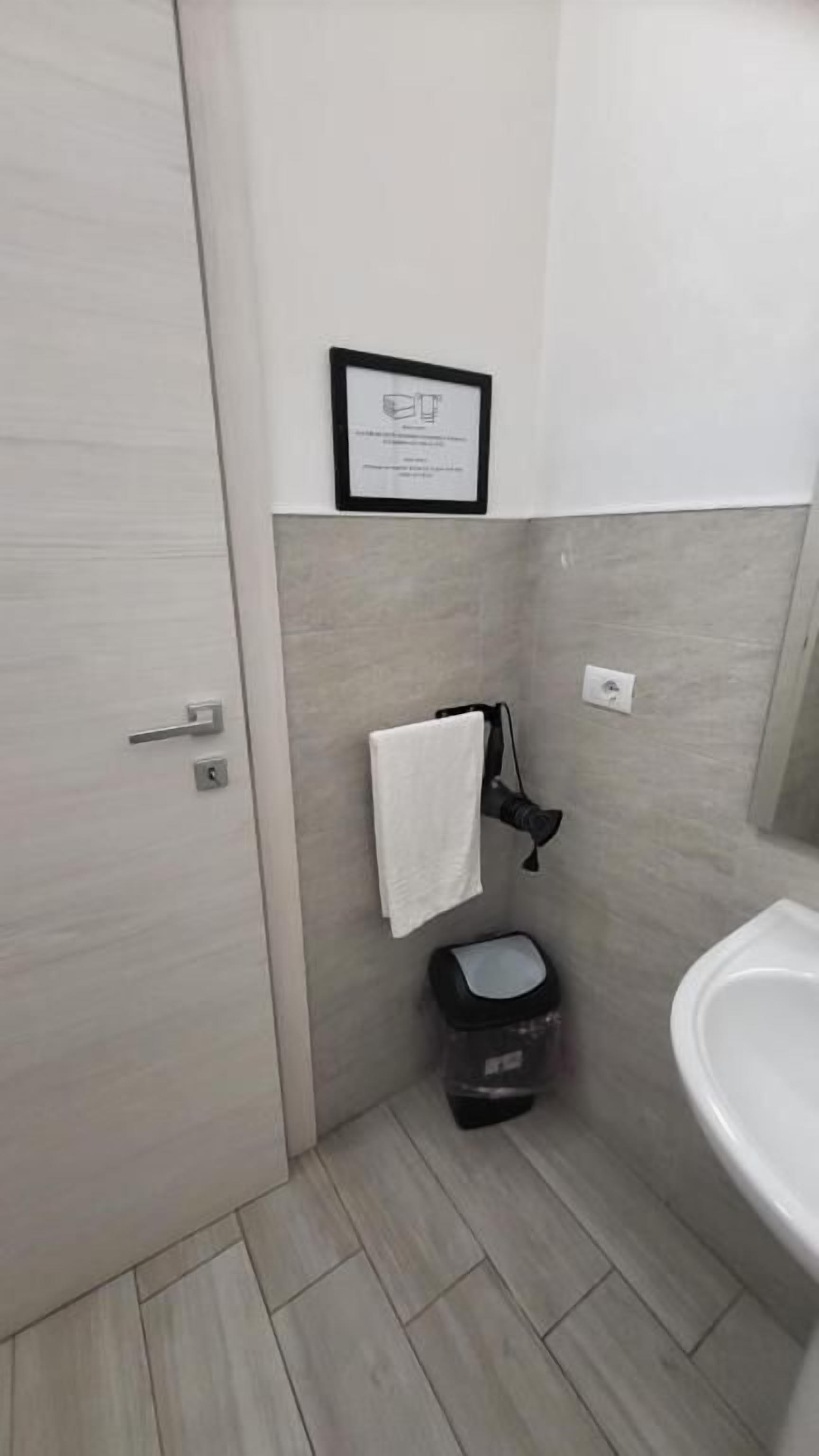 Et lille badeværelse med toilet, håndvask, et håndklæde hængende på en krog og et indrammet certifikat på væggen.