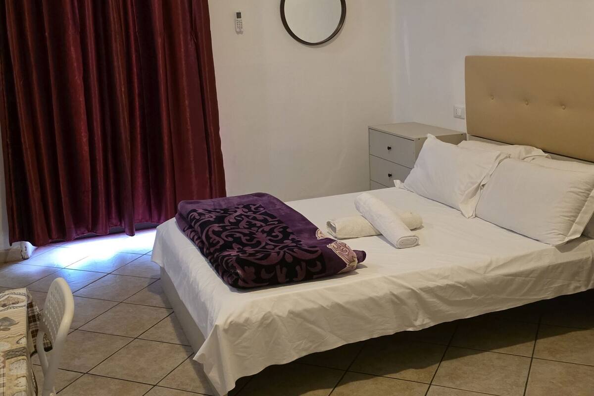 A bedroom with a bed, a bedside table, a chair, a curtain, and an air conditioning unit.