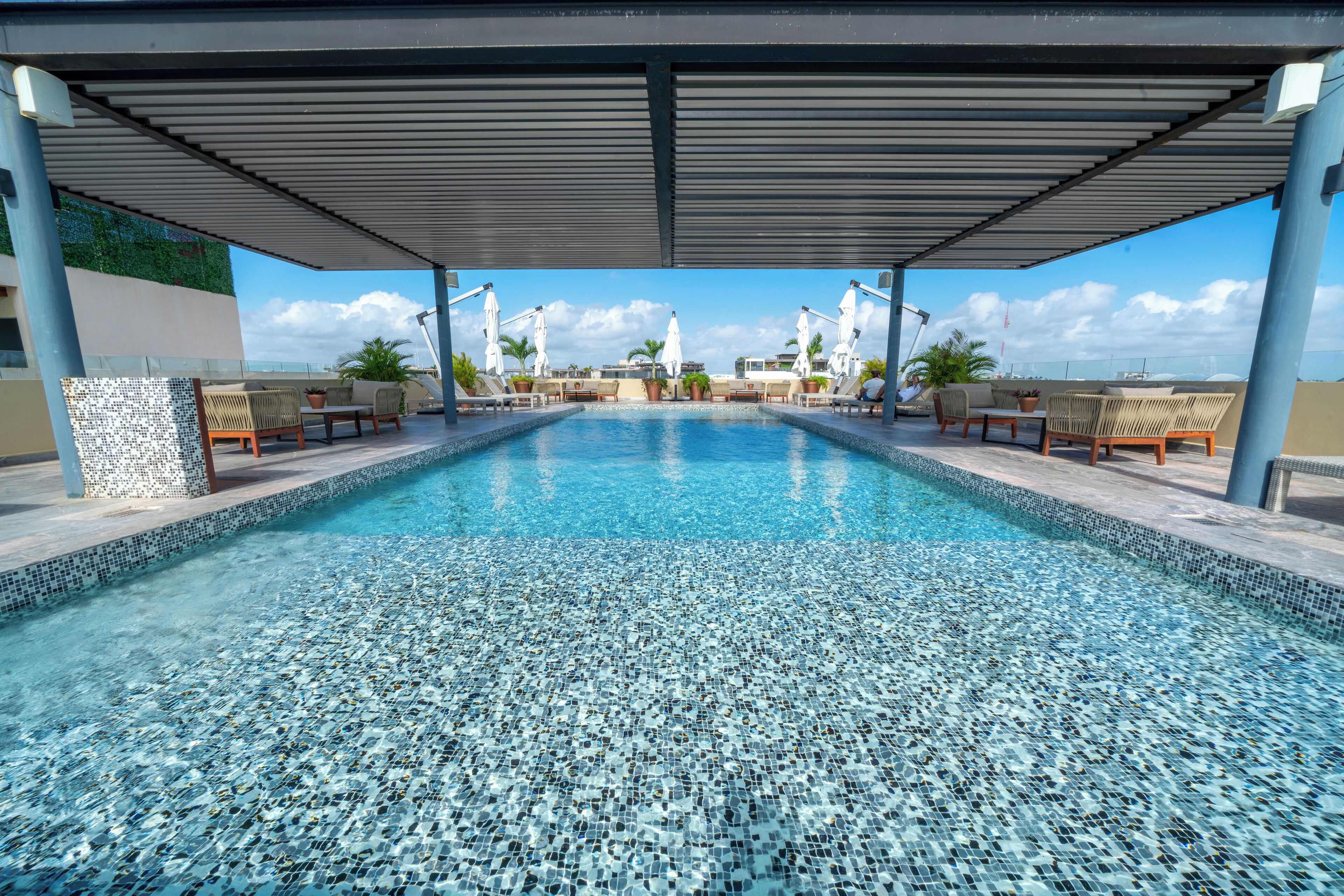 A pool with mosaic tiles, lounge chairs, and umbrellas under a covered area.