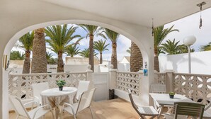A covered outdoor seating area with white chairs and tables, palm trees, and a white wall in the background.