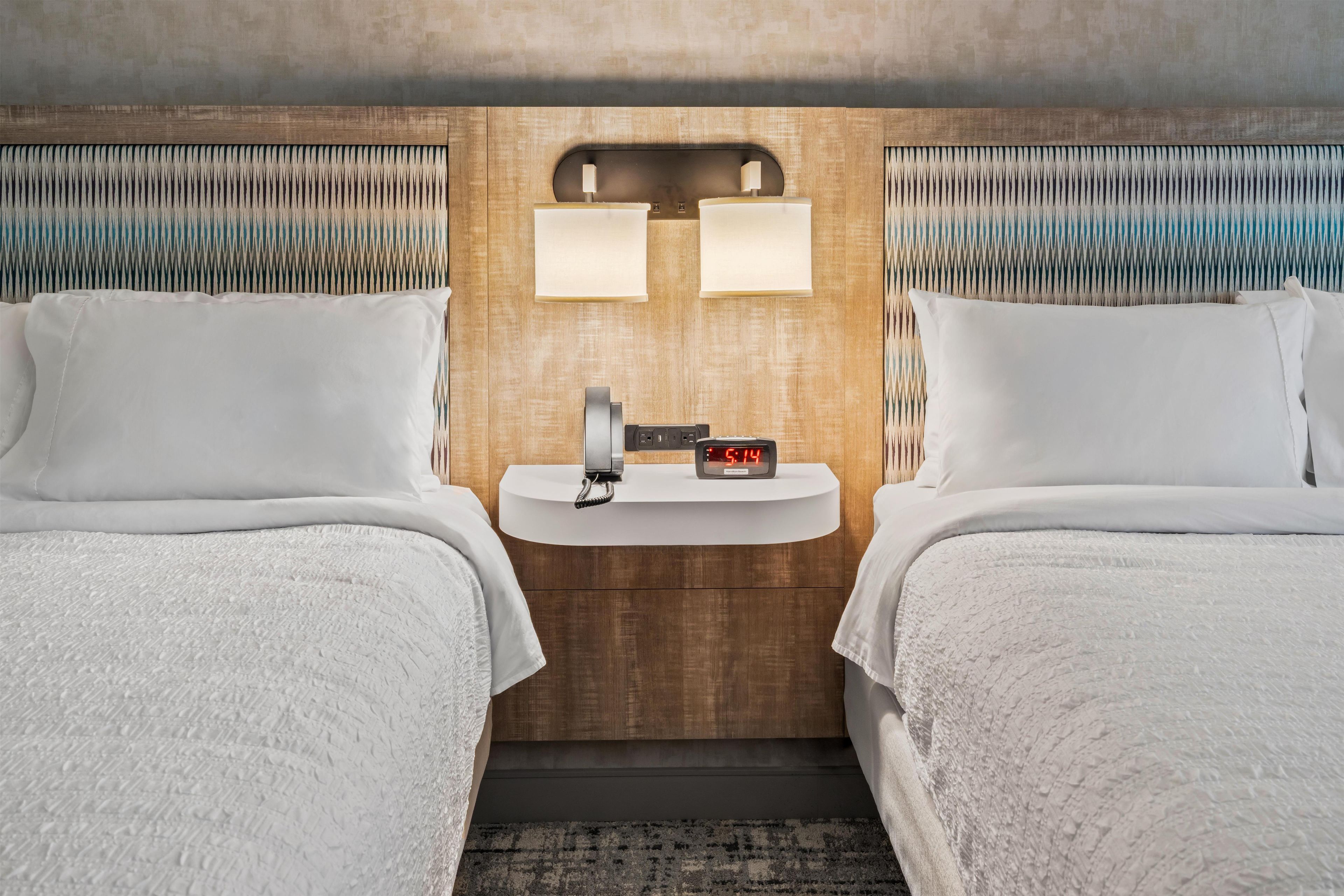 A hotel room with two beds, a bedside table with a clock, and a wall-mounted light fixture.