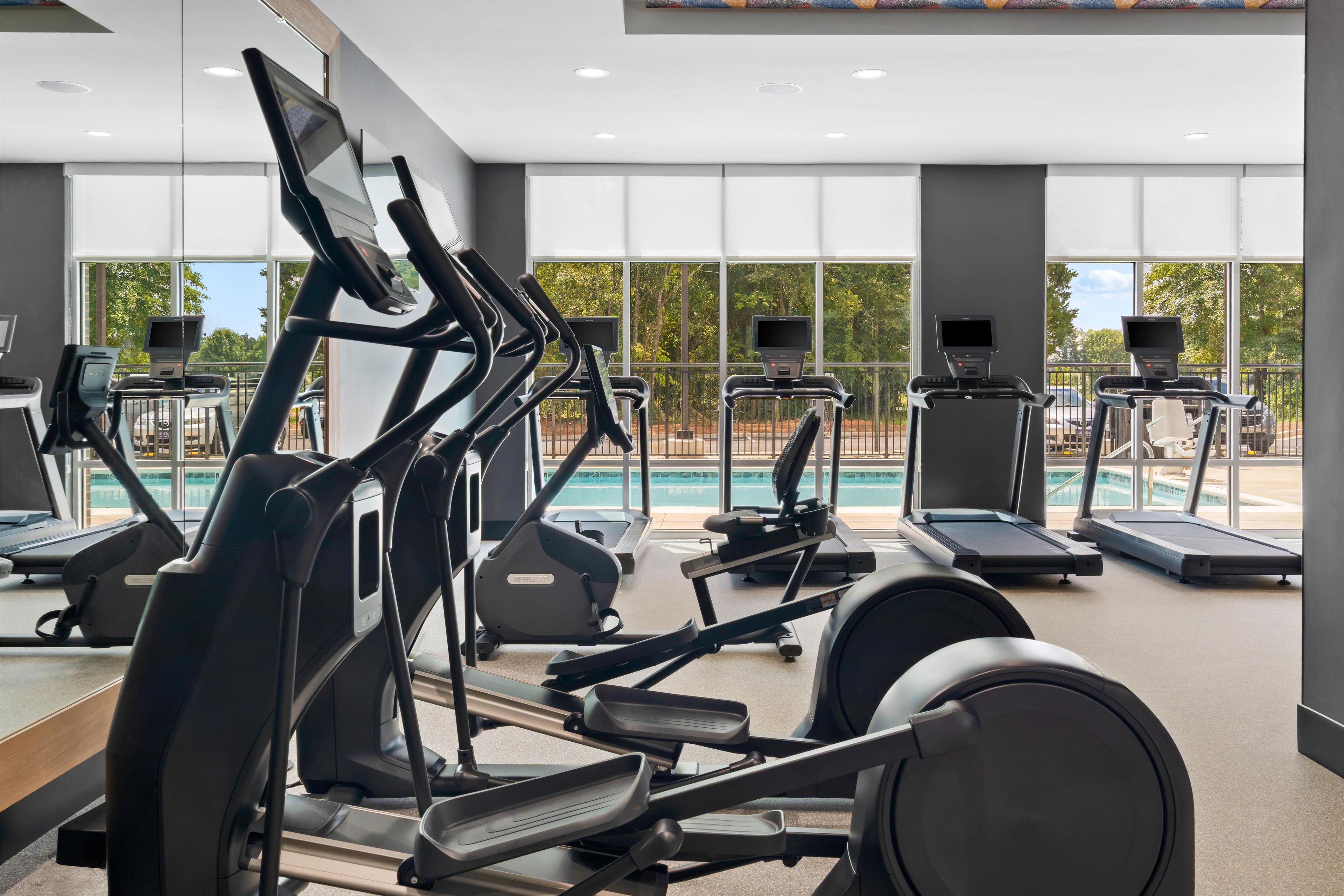 A gym with multiple exercise machines, large windows, and a view of a swimming pool.
