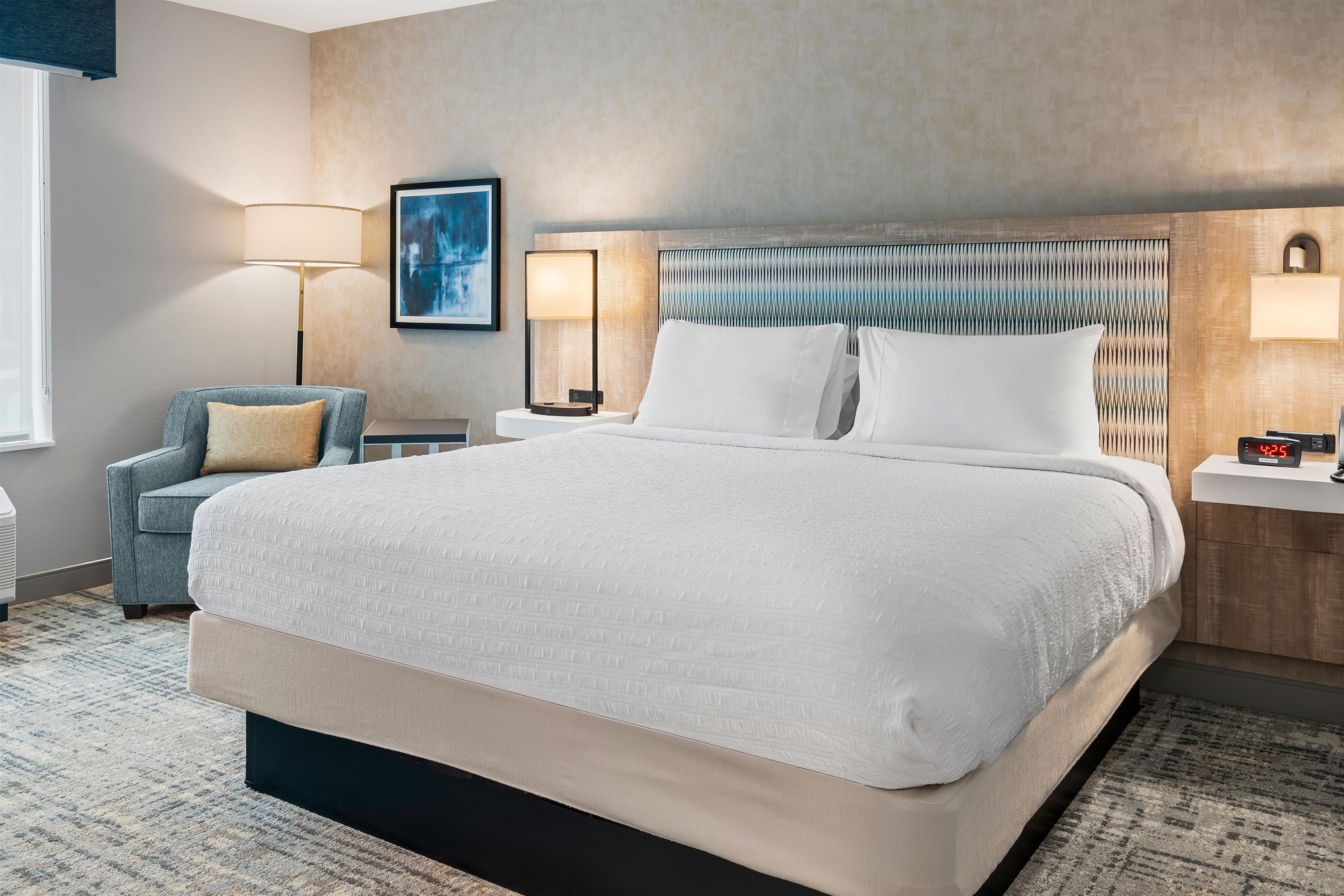 A hotel room with a large bed, a chair, a bedside table, a lamp, and a wall-mounted artwork.