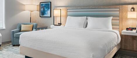 Habitación de hotel con una cama grande, una silla, una mesita de noche, una lámpara y una obra de arte colgada en la pared.