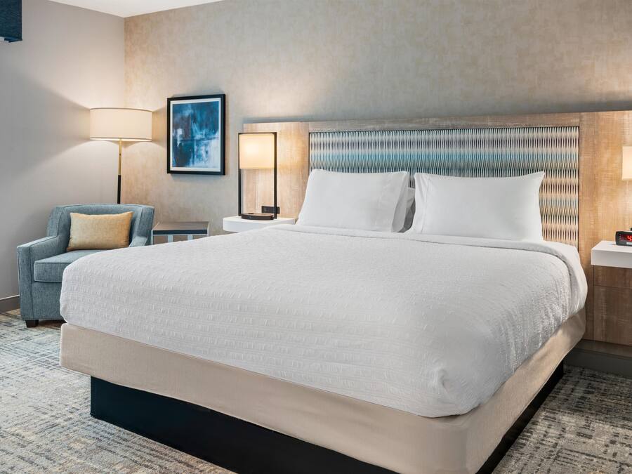 A hotel room with a large bed, a chair, a bedside table, a lamp, and a wall-mounted artwork.