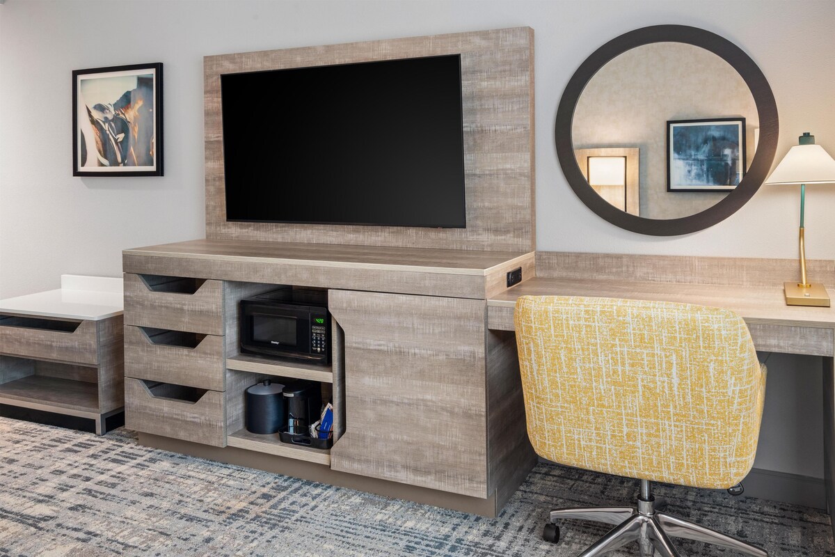 A hotel room with a flat-screen TV mounted on a wooden cabinet, a microwave, a coffee maker, and a chair with a yellow patterned cushion.