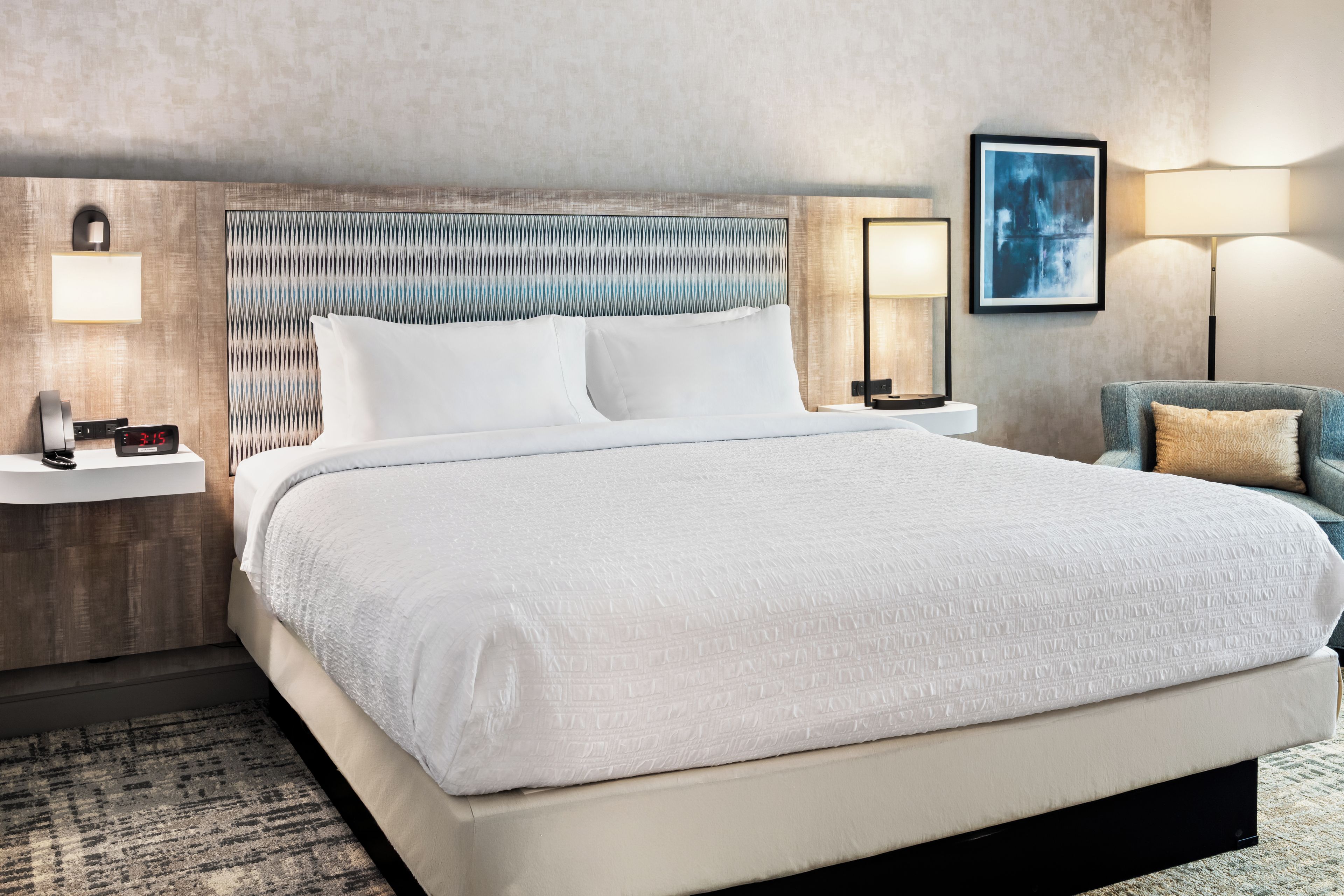 A hotel room with a large bed, a nightstand with a lamp, a wall-mounted clock, and a framed picture on the wall.