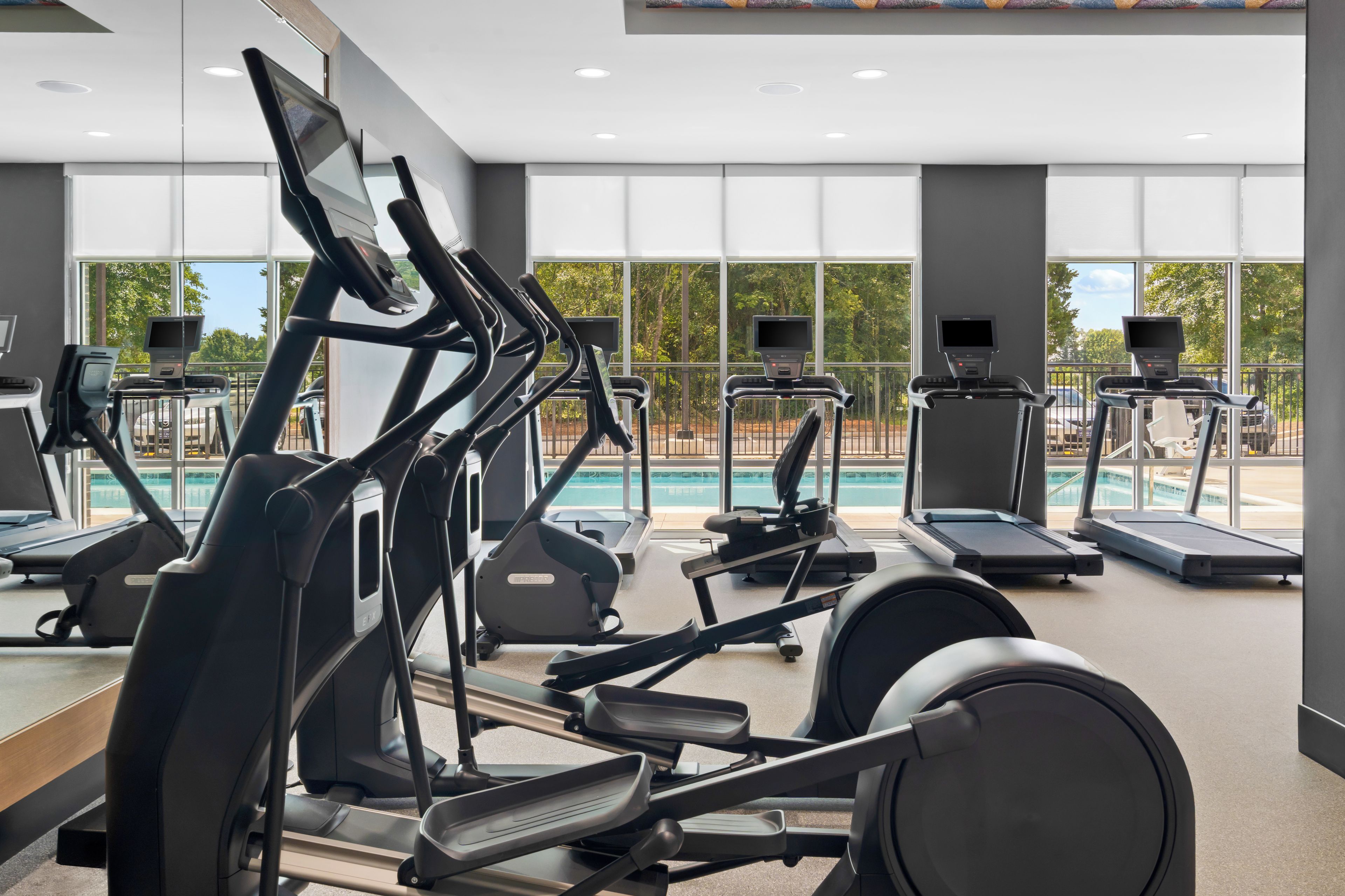 A gym with multiple exercise machines, large windows, and a view of a swimming pool.
