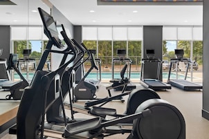 Academia com vários equipamentos de exercício, grandes janelas e vista para uma piscina.