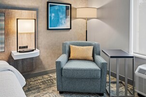 Quarto de hotel com uma poltrona cinza, luminária de pé, quadro emoldurado e uma mesinha.