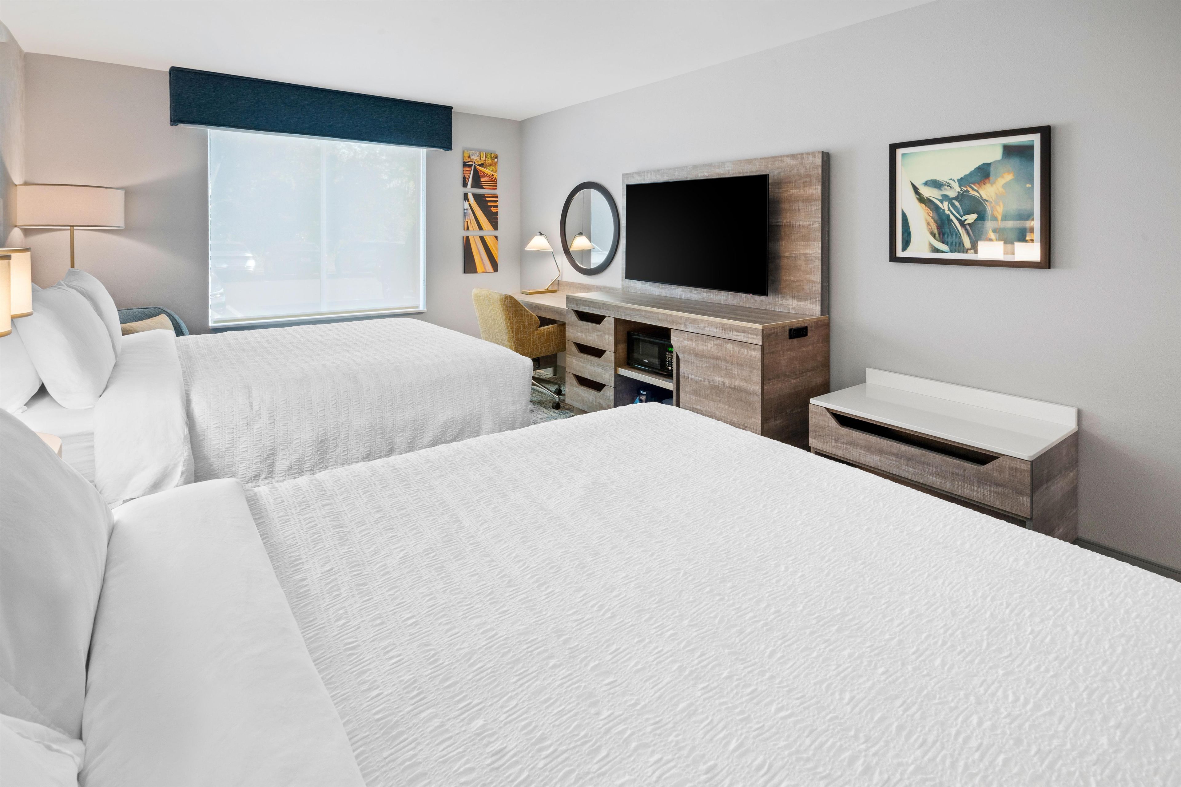 A hotel room with two beds, a desk, a flat-screen TV, a mirror, and artwork on the walls.