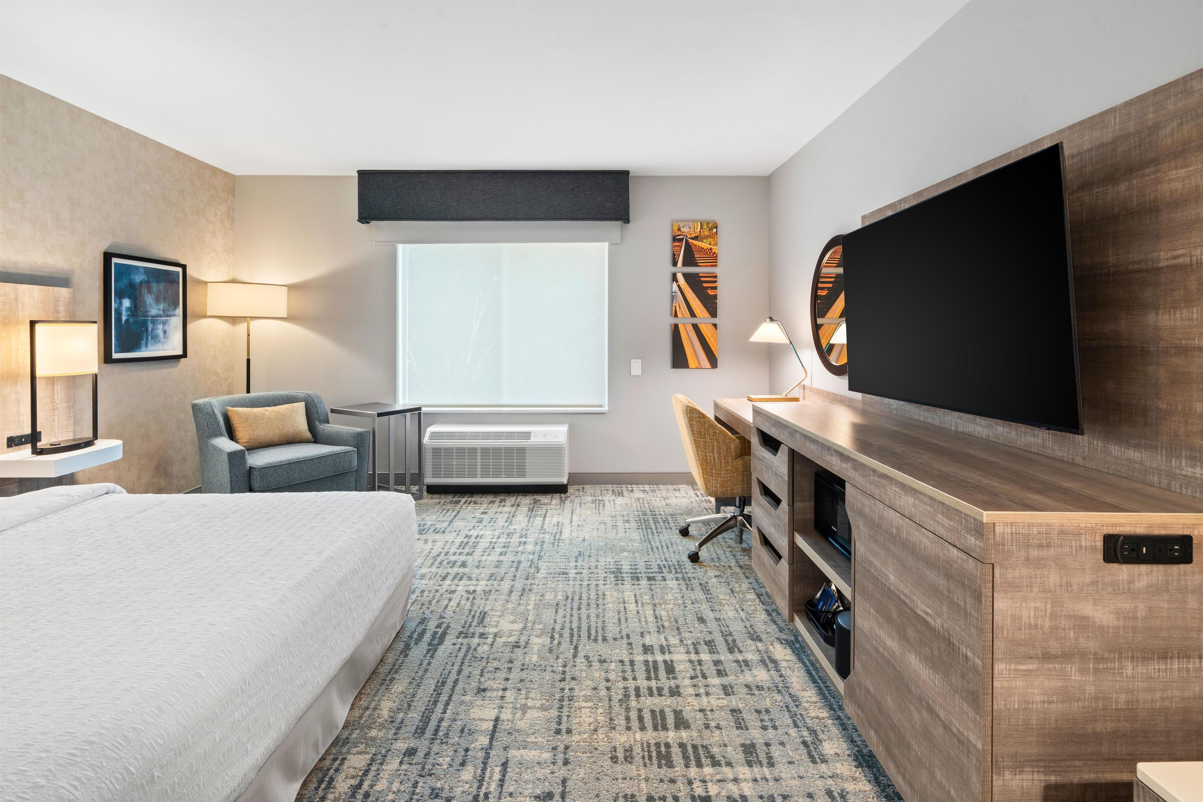 A hotel room with a large bed, a desk with a flat-screen TV, a chair, a lamp, and a window.