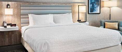 A hotel room with a large bed, a headboard with a textured pattern, bedside tables with lamps, a framed picture on the wall, and a blue armchair.