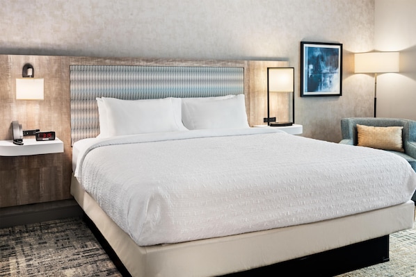 Una habitación de hotel con una cama grande, cabecera con diseño texturizado, mesitas de noche con lámparas, un cuadro enmarcado en la pared y un sillón azul.