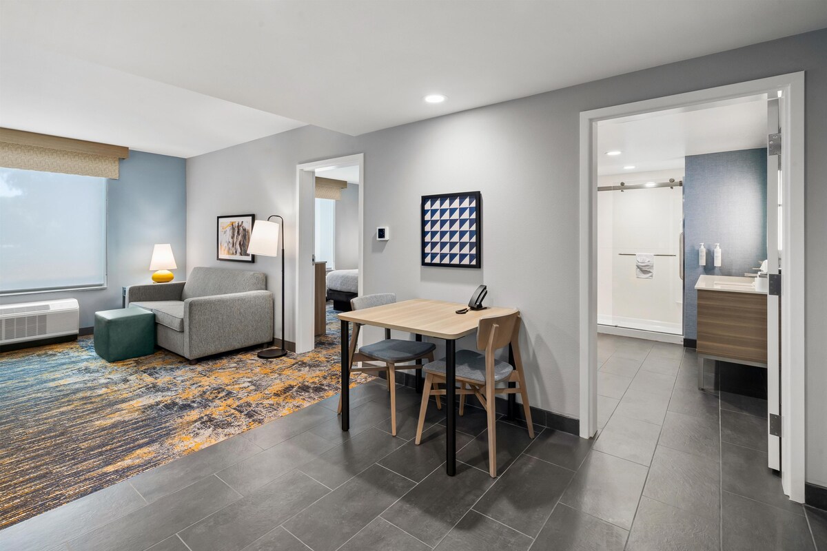 Habitación de hotel moderna con comedor, un sofá gris, un tapete estampado y un baño visible a través de una puerta abierta.
