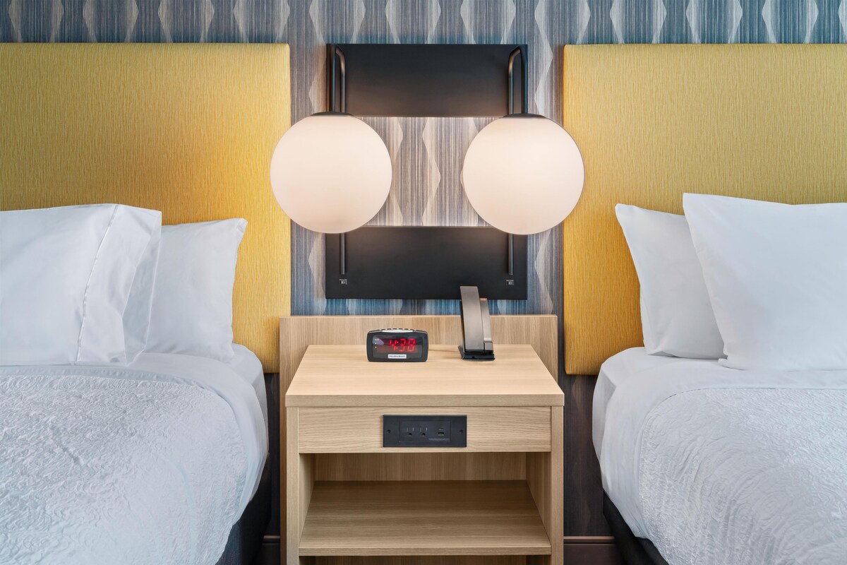 Habitación de hotel con dos camas, una mesita de noche con un reloj digital y un televisor de pared.