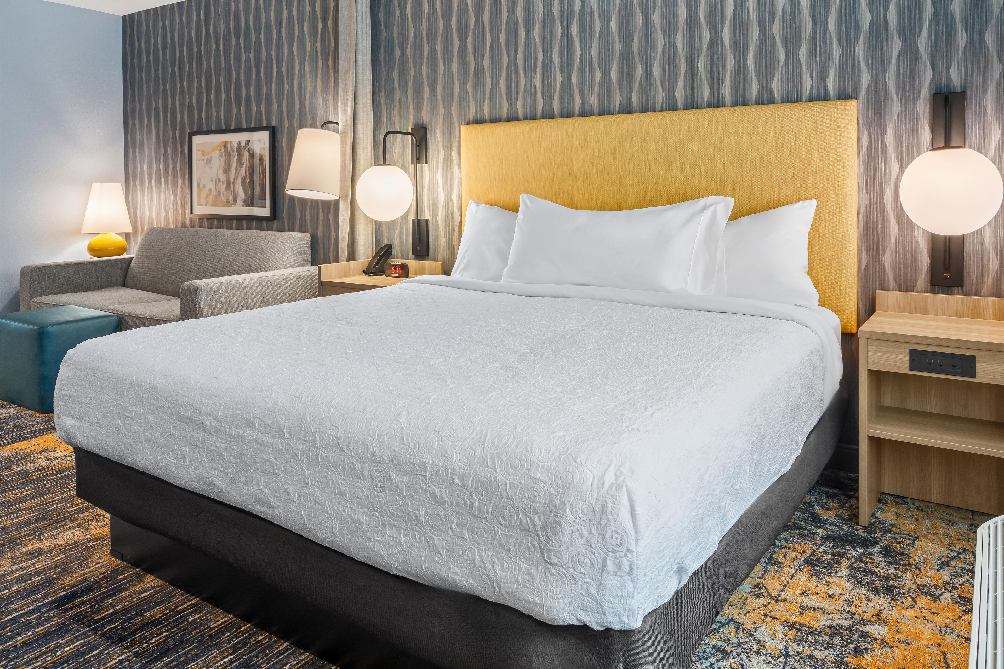 A hotel room with a large bed, a bedside table, a chair, a lamp, and a wall-mounted phone.