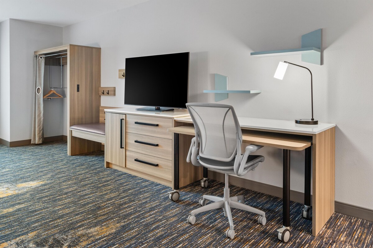 Habitación de hotel con escritorio de madera, una silla moderna, televisor de pantalla plana, lámpara de escritorio y armario con espejo.
