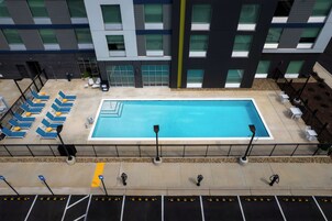 Una piscina con tumbonas y una zona de estar cubierta, rodeada por un edificio con grandes ventanales y un paseo empedrado.
