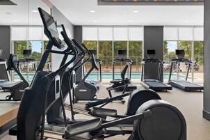 Un gimnasio con varias máquinas de ejercicio, ventanas amplias y vistas a una piscina.
