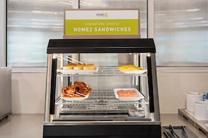 Presentación de alimentos con sándwiches y tocino, con el rótulo 'Signature Choice Home2 Sandwiches'.