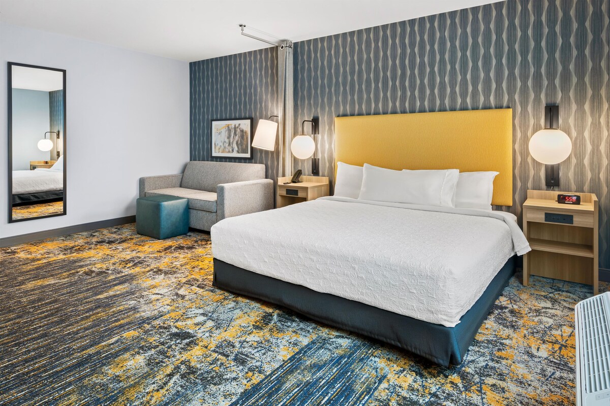 Habitación de hotel con una cama grande, un escritorio, una silla y una mesita de noche con lámpara.