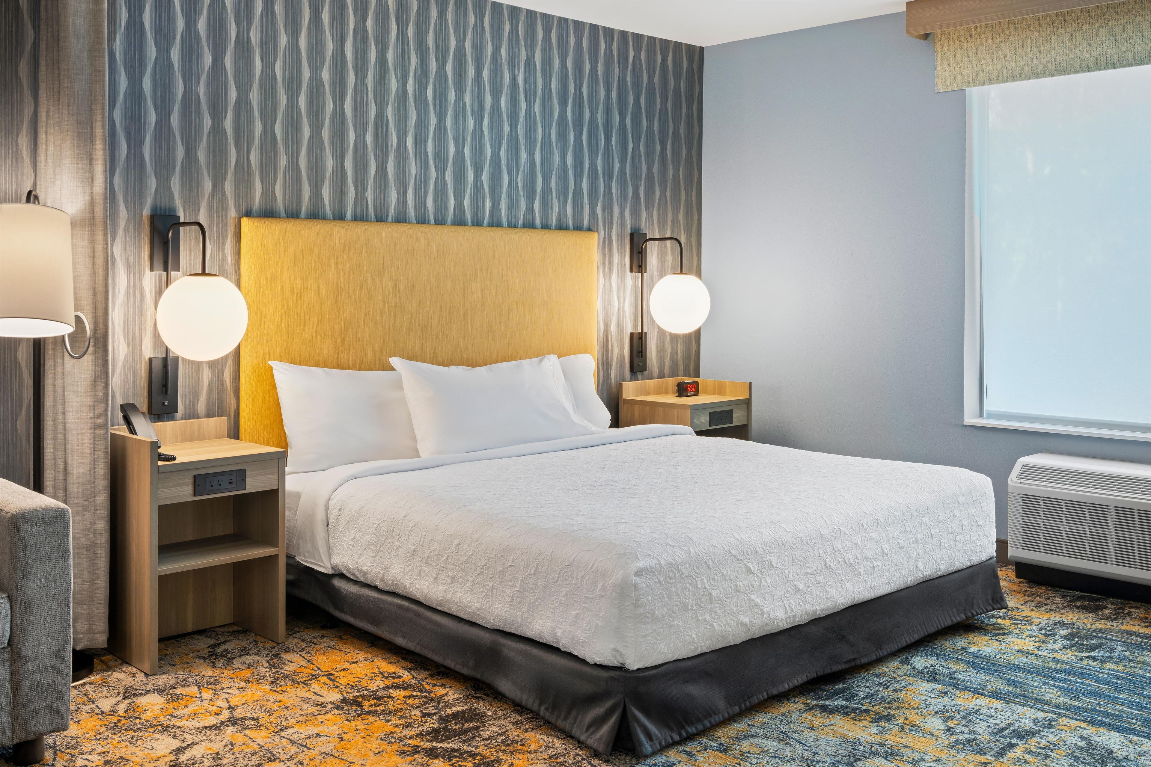 Una habitación de hotel con una cama grande, mesitas de noche, una lámpara, un reloj y una ventana.