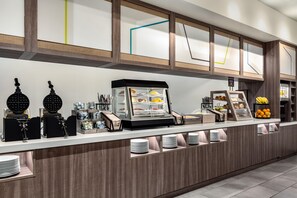 Een moderne ontbijtbar met een koffieapparaat, een vitrine met gebak en een fruitschaal.