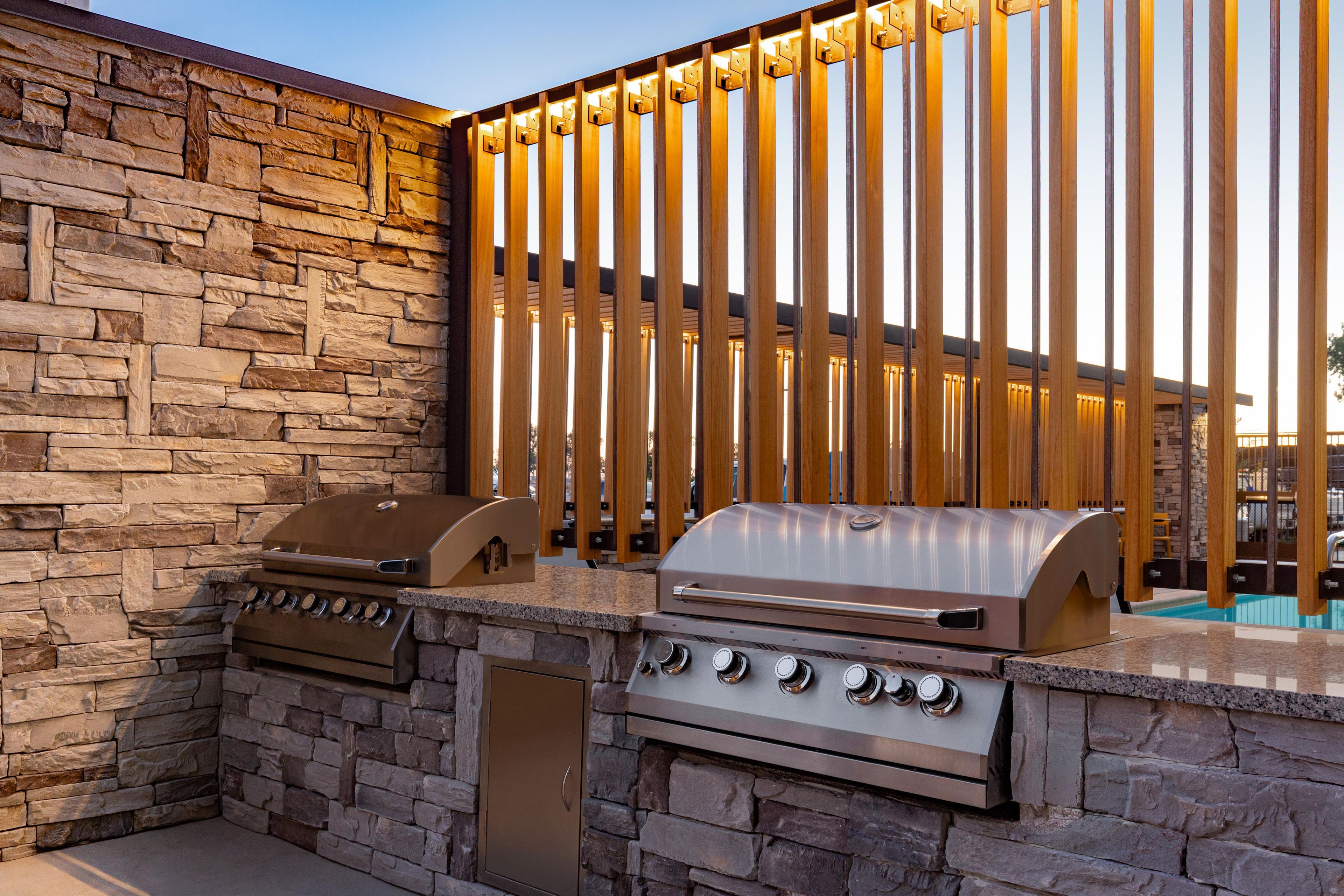 An outdoor kitchen with a stone wall, two grills, and a wooden fence with lights.