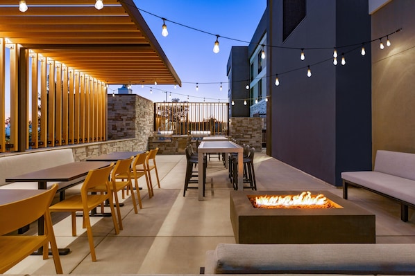 Un patio al aire libre con mesas, sillas, un hogar para fuego y luces colgantes.