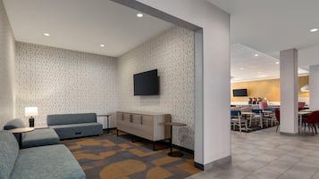 Un moderno vestíbulo de hotel con una zona de estar, un televisor de pantalla plana en la pared y un área de comedor al fondo.