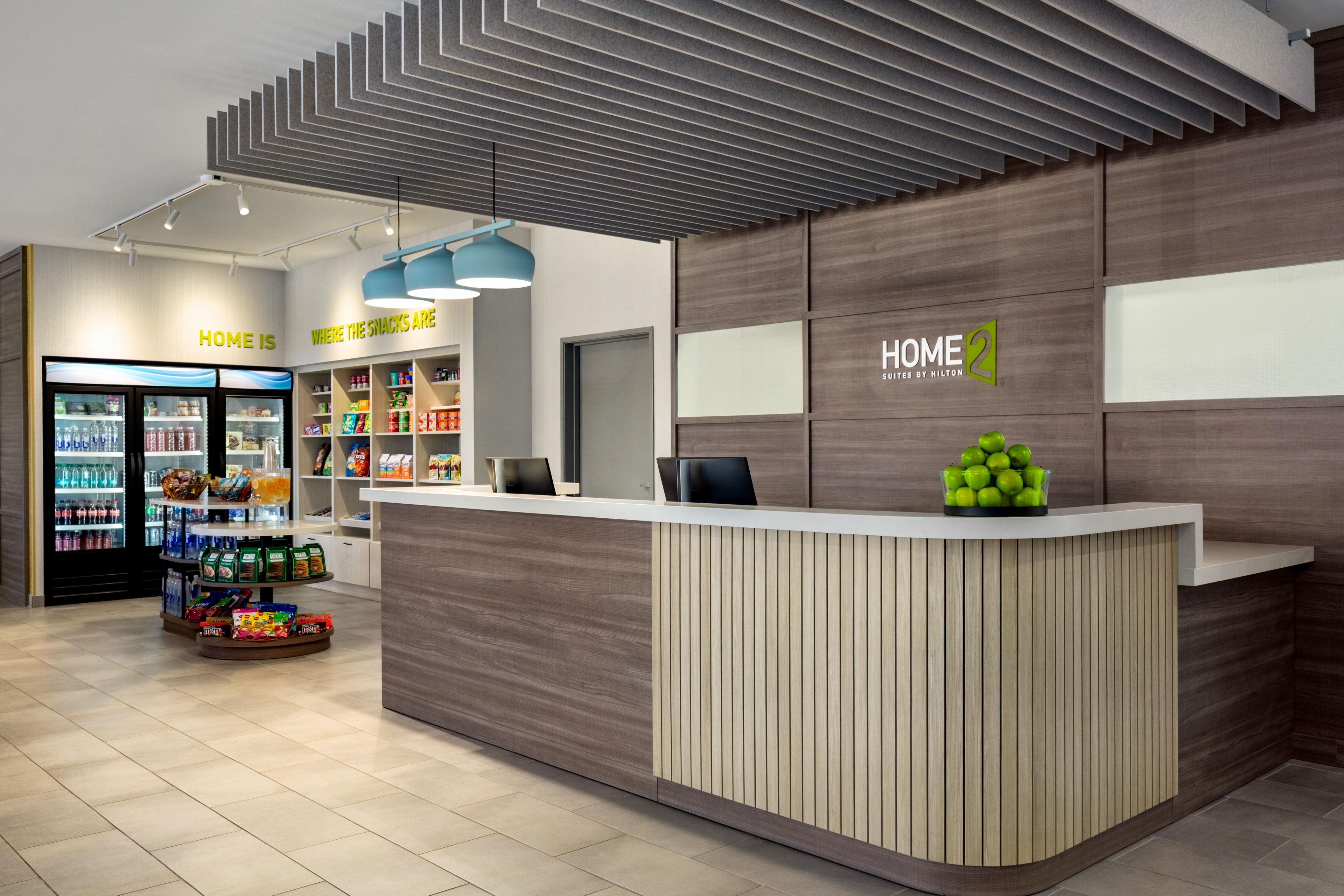 A modern hotel reception area with a wooden reception desk, a snack bar in the background, and a fruit display.