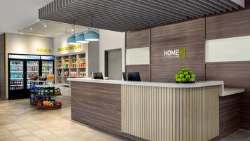A modern hotel reception area with a wooden reception desk, a snack bar in the background, and a fruit display.