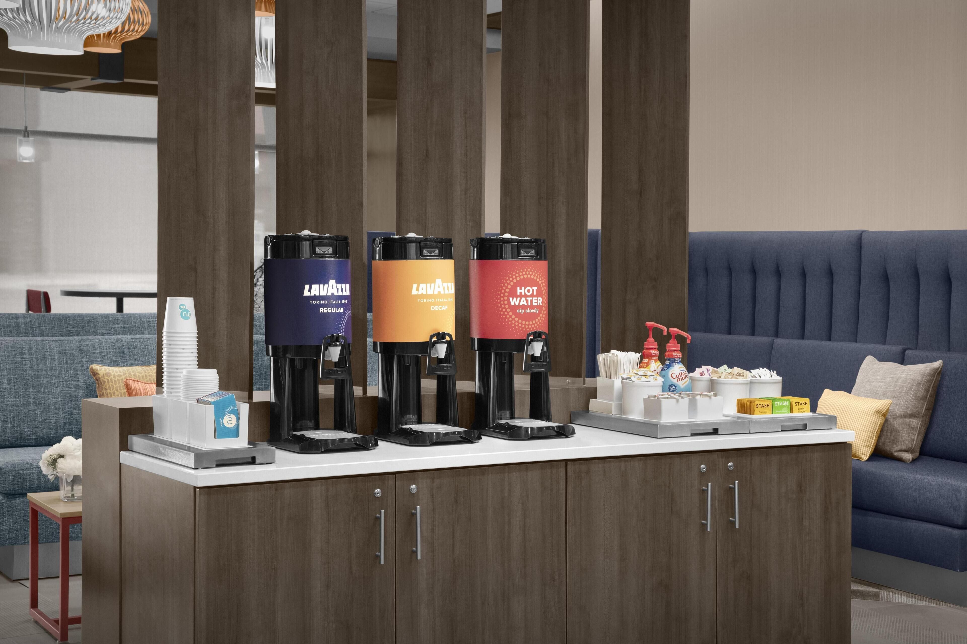 A coffee station with three coffee makers, a water dispenser, and a snack bar with various items.