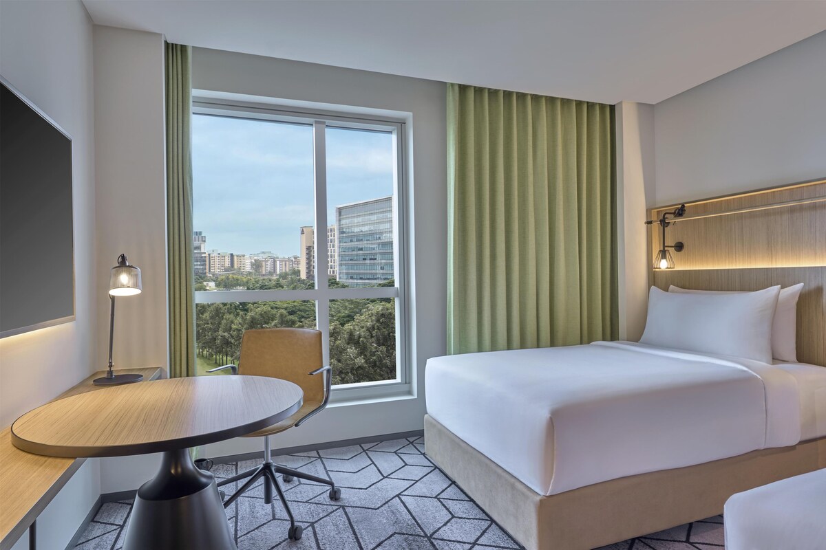 A hotel room with a bed, a desk, a chair, a window with curtains, and a view of buildings.