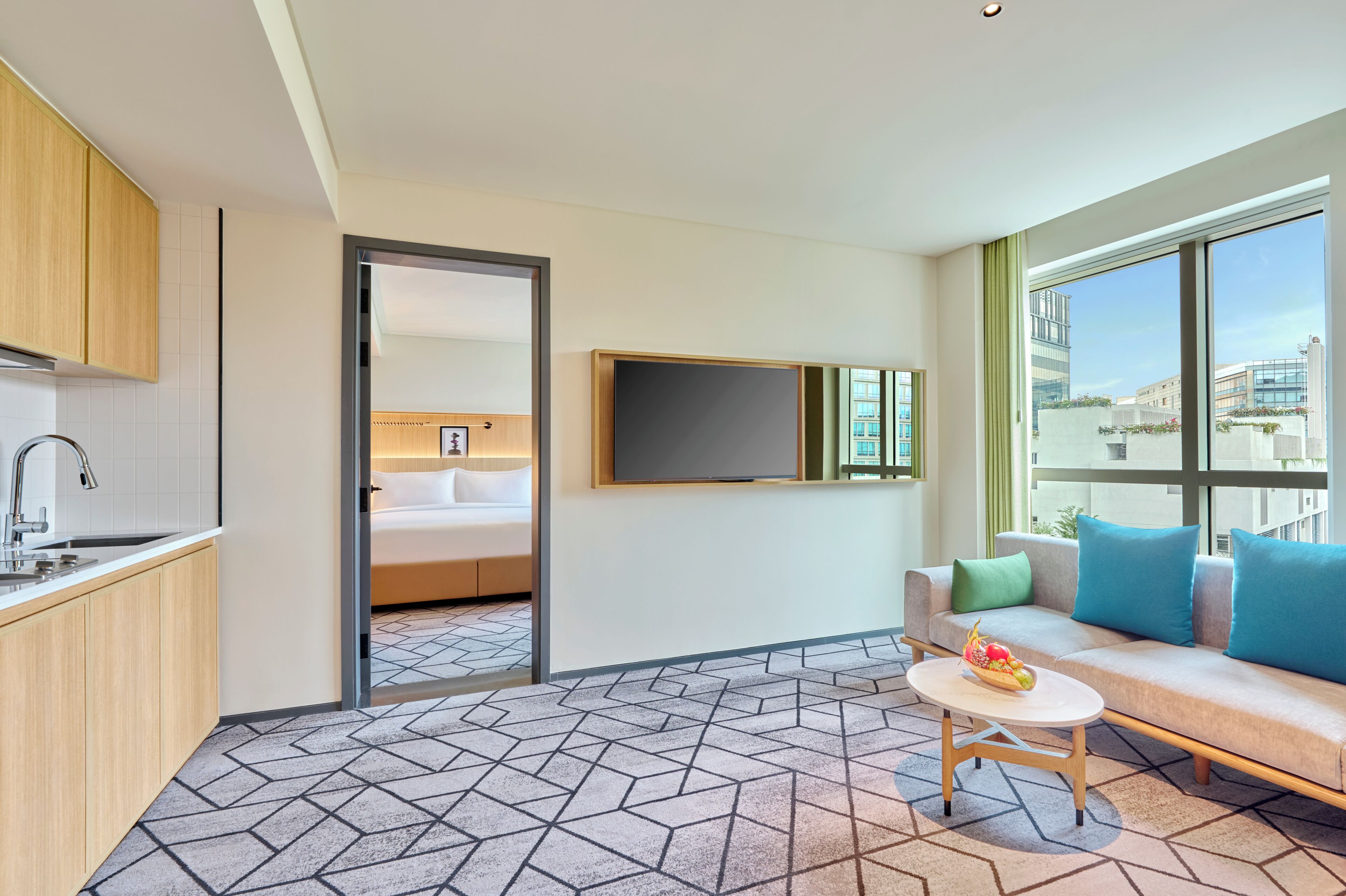 Una habitación de hotel moderna con kitchenette, una cama con cabecera, un televisor montado en la pared, un sofá con cojines y una ventana con vista a la ciudad.