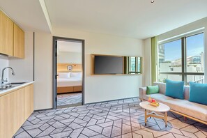 A modern hotel room with a kitchenette, a bed with a headboard, a TV mounted on the wall, a sofa with cushions, and a window with a city view. - Hilton Garden Inn Bengaluru Embassy TechVillage Outer Ring Road (Bengaluru)