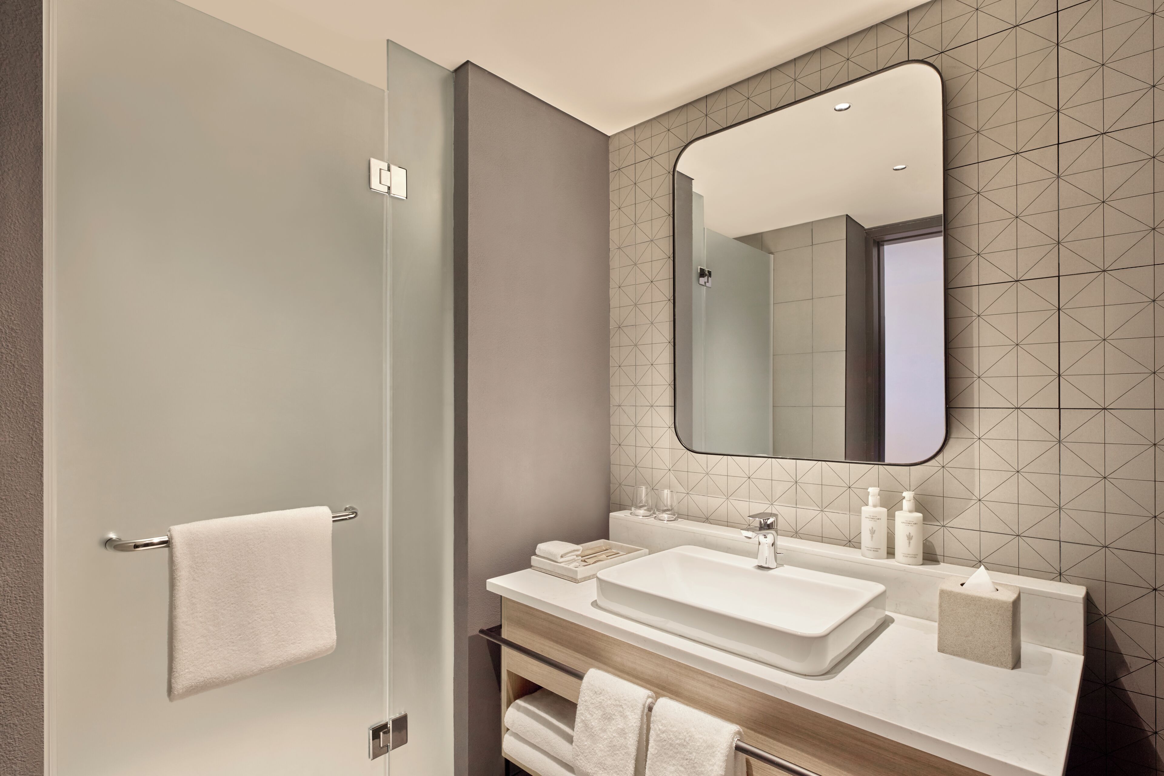 Un baño moderno con puerta de ducha de vidrio, un espejo grande, un lavamanos blanco y un mueble de tocador con artículos de aseo.