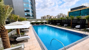 A rectangular swimming pool with a metal ladder, surrounded by lounge chairs and umbrellas, with modern buildings in the background. - Hilton Garden Inn Bengaluru Embassy TechVillage Outer Ring Road (Bengaluru)