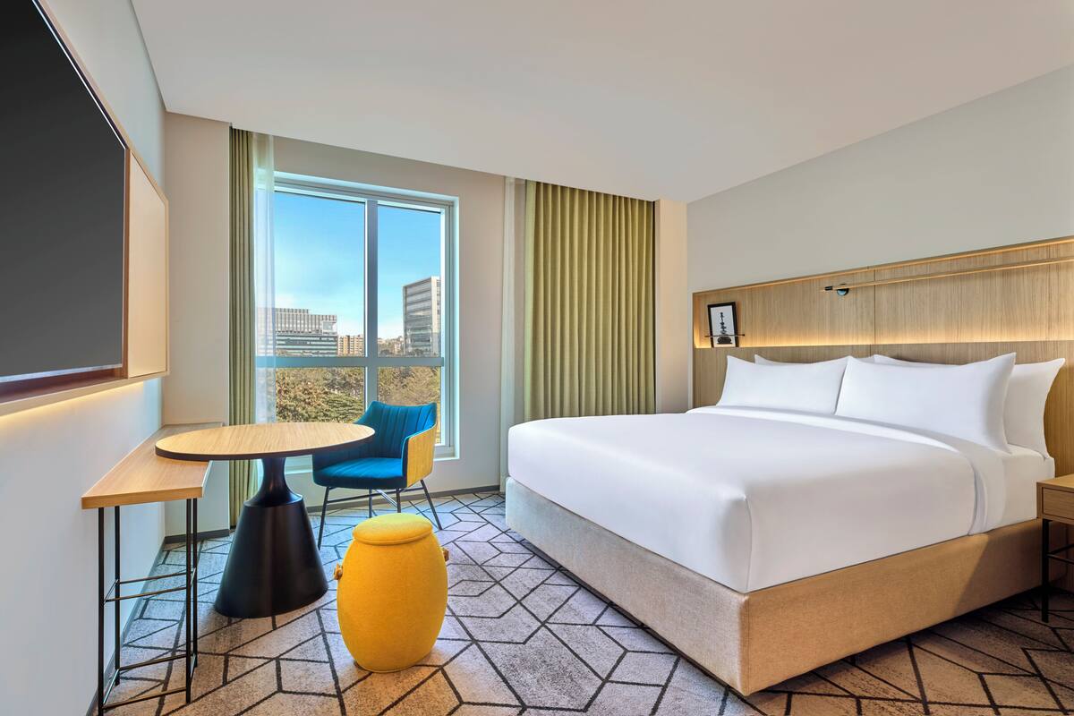 Habitación de hotel con cama, una mesa, una silla, un reposapiés amarillo y un ventanal con vista a la ciudad.