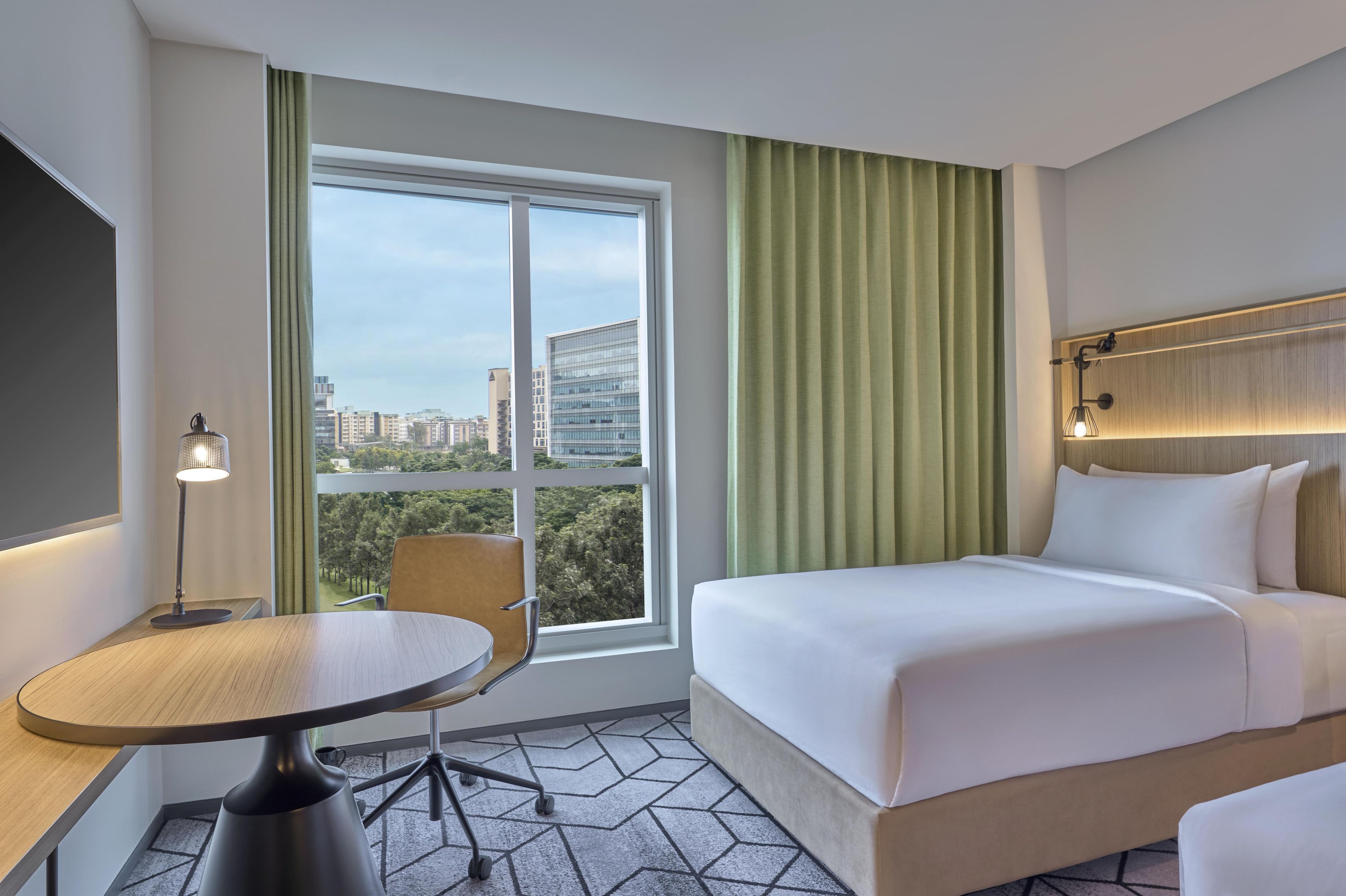A hotel room with a bed, a desk, a chair, a window with curtains, and a view of buildings.